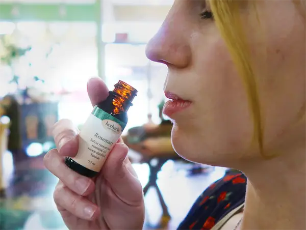 A close-up side profile of a woman holding an open small amber glass bottle of rosemary essential oil toward her nose to smell it.