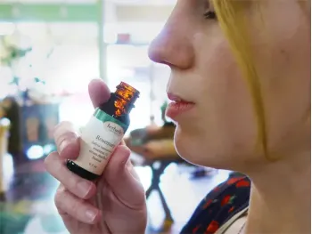A close-up side profile of a woman holding an open small amber glass bottle of rosemary essential oil toward her nose to smell it.