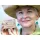 A smiling woman in a straw hat holding a bar of Herbaria all-natural Lavender Oatmeal Soap. She wears green teardrop earrings that fit the natural, earthy aesthetic of the brand.