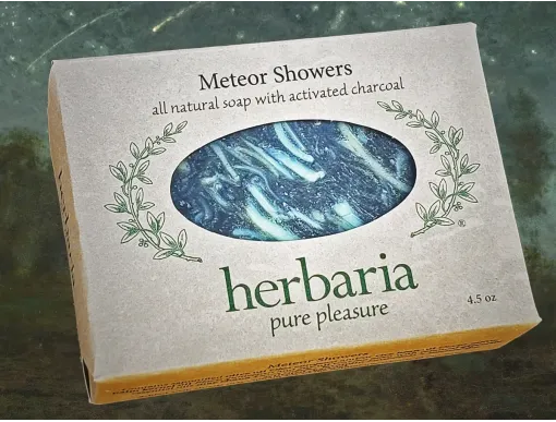 A bar of Herbaria soap Meteor Showers wrapped in a paper label that showcases it's colorful scraps in a black bar on a background of a meteor shower.