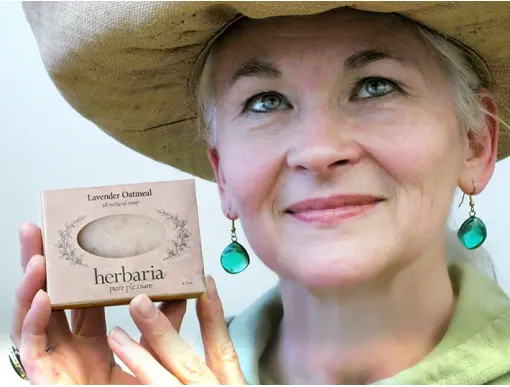 A smiling woman in a straw hat holding a bar of Herbaria all-natural Lavender Oatmeal Soap. She wears green teardrop earrings that fit the natural, earthy aesthetic of the brand.