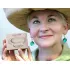 A smiling woman in a straw hat holding a bar of Herbaria all-natural Lavender Oatmeal Soap. She wears green teardrop earrings that fit the natural, earthy aesthetic of the brand.