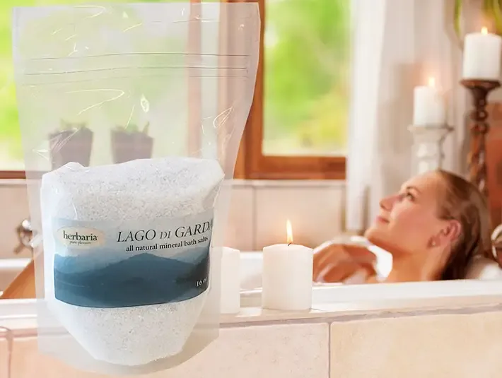 woman bathing in tub with candles and a bigger picture of Herbaria bath salts over top of the image
