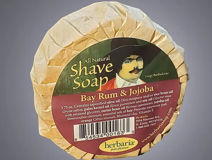 A round bar of Herbaria all-natural Bay Rum shave soap featuring the embossed Herbaria logo on a rustic brown paper wrap.