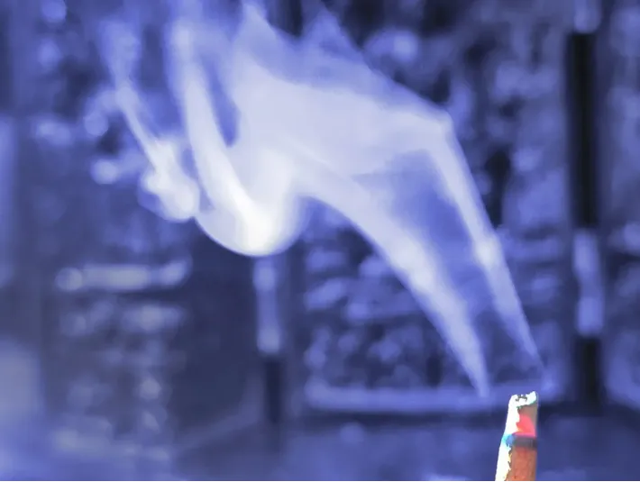 Wisps of blue-toned smoke curling upward from a lit herbal incense stick.