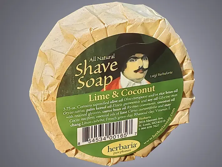 A round bar of Herbaria all-natural Lime & Coocnut shave soap featuring the embossed Herbaria logo on a rustic brown paper wrap.