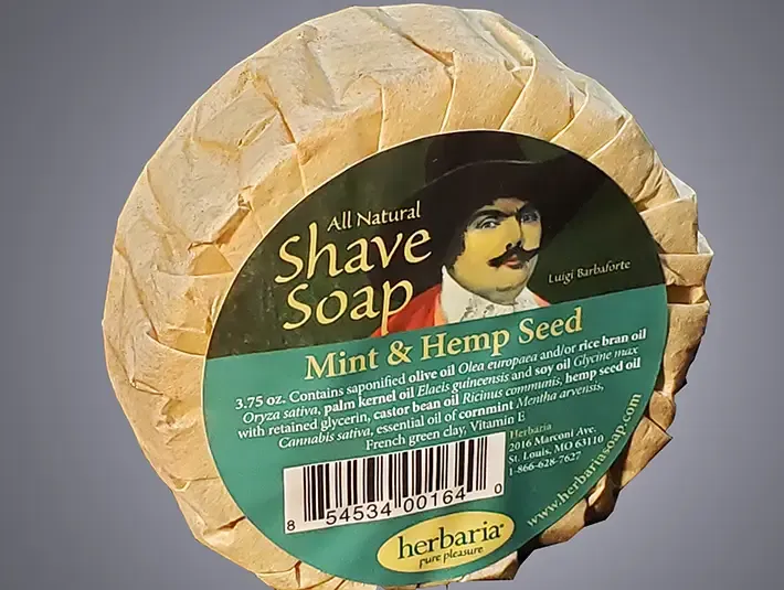 A round bar of Herbaria all-natural Mint & Hemp shave soaps featuring the embossed Herbaria logo on a rustic brown paper wrap.