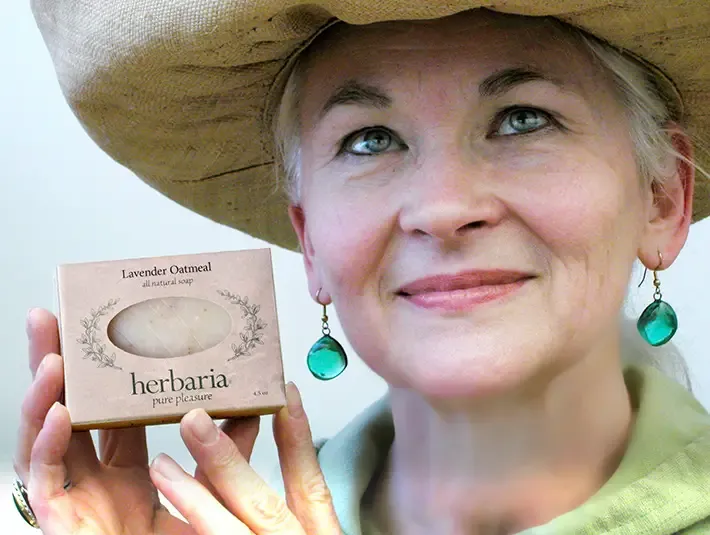 A smiling woman in a straw hat holding a bar of Herbaria all-natural Lavender Oatmeal Soap. She wears green teardrop earrings that fit the natural, earthy aesthetic of the brand.