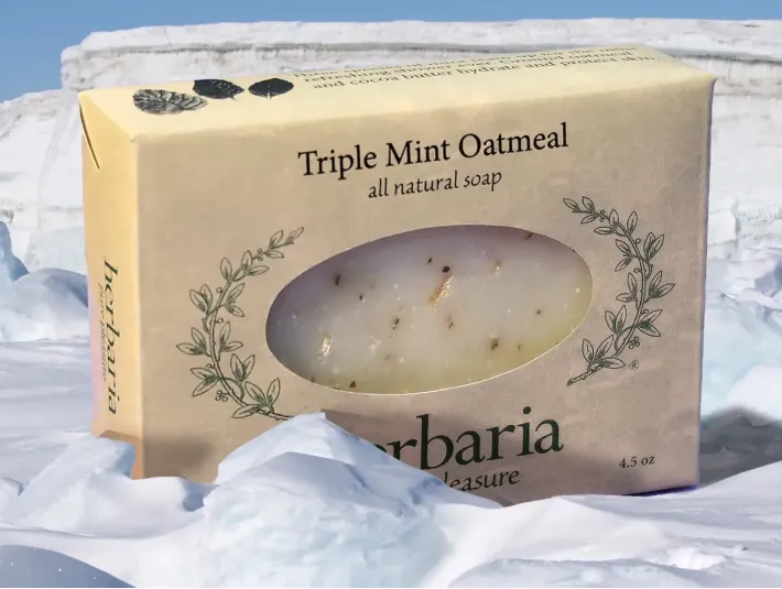 A bar of Herbaria Triple Mint Oatmeal all-natural soap packaged in a tan paper label. The soap bar is visible through an oval window cutout, showing small botanical flecks of real mint leaf inside. The bar is set against a dramatic backdrop of bright white snow and blue-tinted glacial ice blocks, emphasizing a cooling effect.